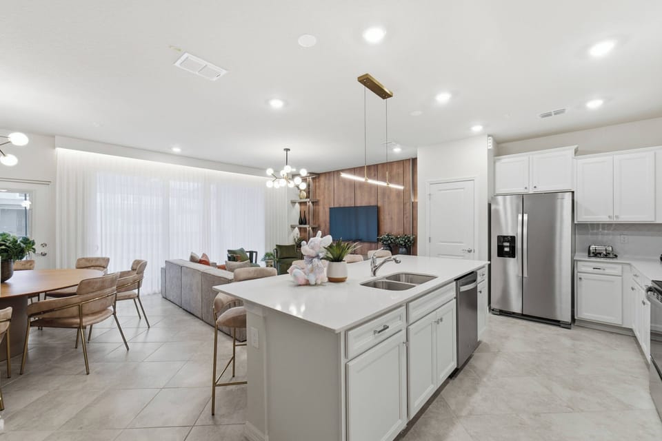 Cook with ease in a bright modern kitchen with white cabinets, stainless steel appliances, spacious counters, a large island, soft lighting, and an open view to the living room, perfect for family meals during vacation.