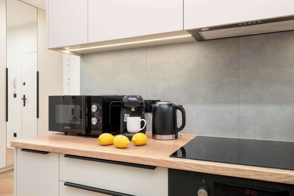 The wooden kitchen countertop holds a black microwave, an electric kettle, and a small bowl containing fresh lemons. Under-cabinet LED lighting provides a warm glow over the workspace.