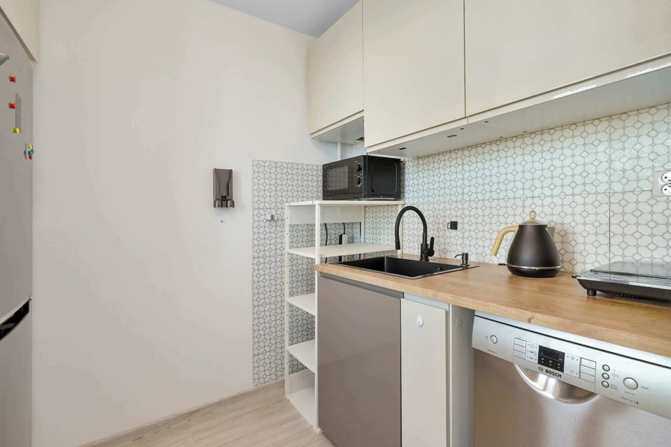 A sleek, minimalist kitchen equipped with light grey cabinetry, a wooden countertop, a modern black sink, and built-in appliances including a dishwasher. A white tiled backsplash complements the contemporary design.