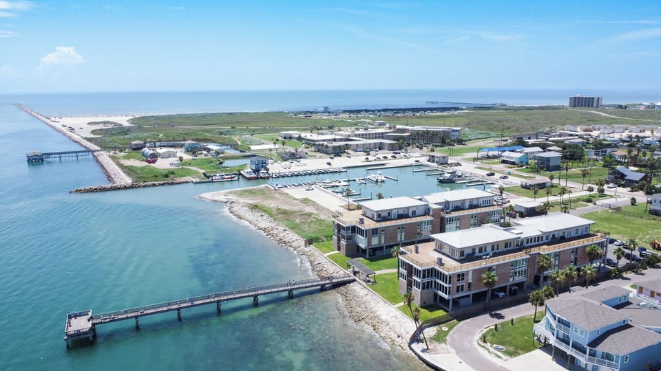 Stunning aerial view of modern waterfront buildings nestled along pristine coastline with marina, pier, and crystal-clear waters stretching to the horizon.
