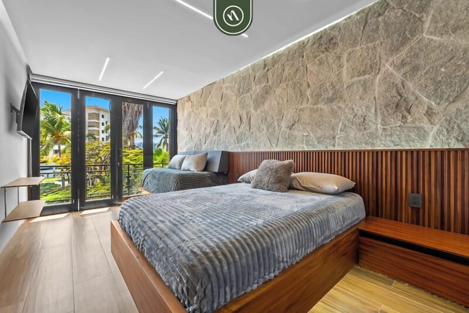 Spacious bedroom with two comfortable beds, warm wood finishes, and a textured stone accent wall. Floor-to-ceiling glass doors bring in natural light and open to a private balcony with tropical views.