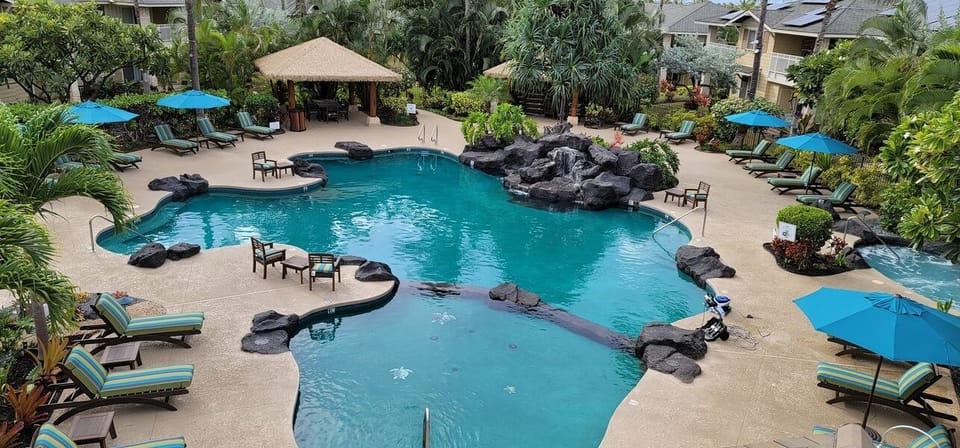 Aerial view of a resort-style lagoon pool surrounded by lush tropical landscaping, lounge seating, bright blue umbrellas, and a central tiki-style pavilion.