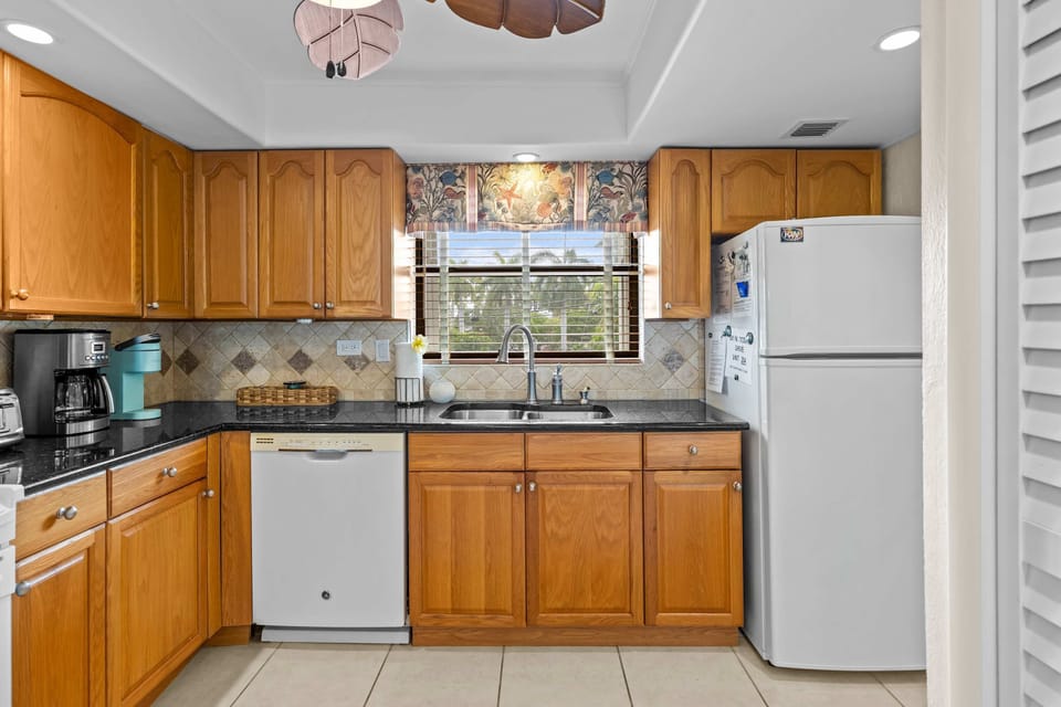 Kitchen Workspace: A window above the sink frames the palms outside while granite counters, a full-size fridge, and a coffee station make morning routines feel unhurried.