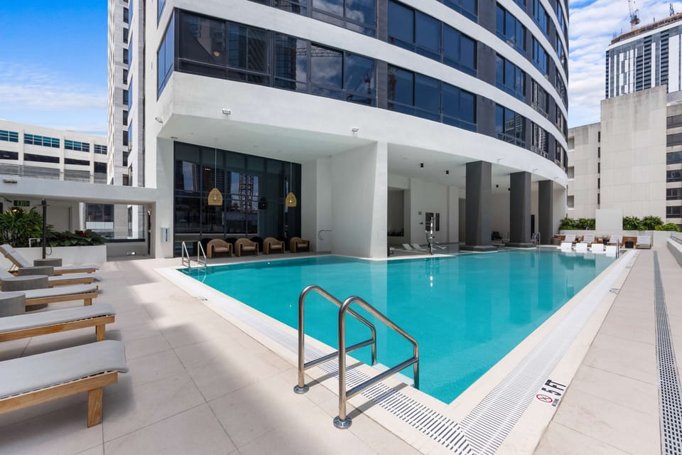 Shared Building Amenity — Resort Pool: A wide turquoise pool with teak lounge chairs runs the length of the building, framed by curving towers and ready for a real swim or a slow afternoon.