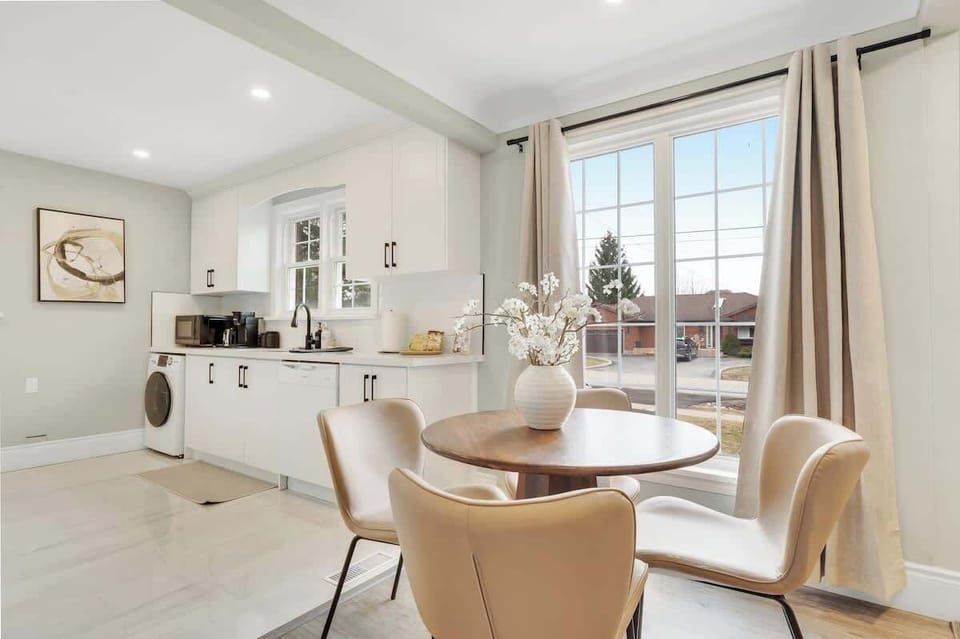Bright and spacious kitchen and dining area featuring a round wooden table with four cushioned chairs, large window with flowing curtains, modern appliances, coffee maker, and washer.