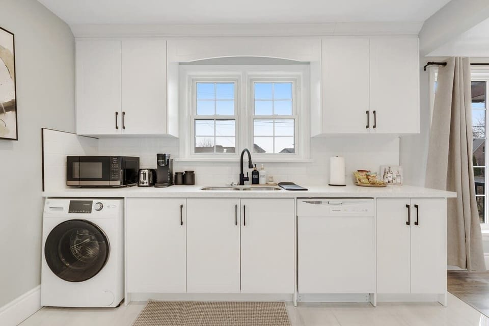 Modern kitchen with sleek white cabinetry, a front-load washer, microwave, coffee maker, blender, toaster, dishwasher, double windows, and a spacious sink area.