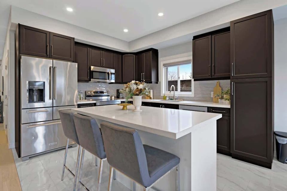 Sleek modern kitchen with spacious white island, three cushioned bar stools, stainless steel fridge, microwave, oven, sink, stylish cabinetry, and floral centerpiece.