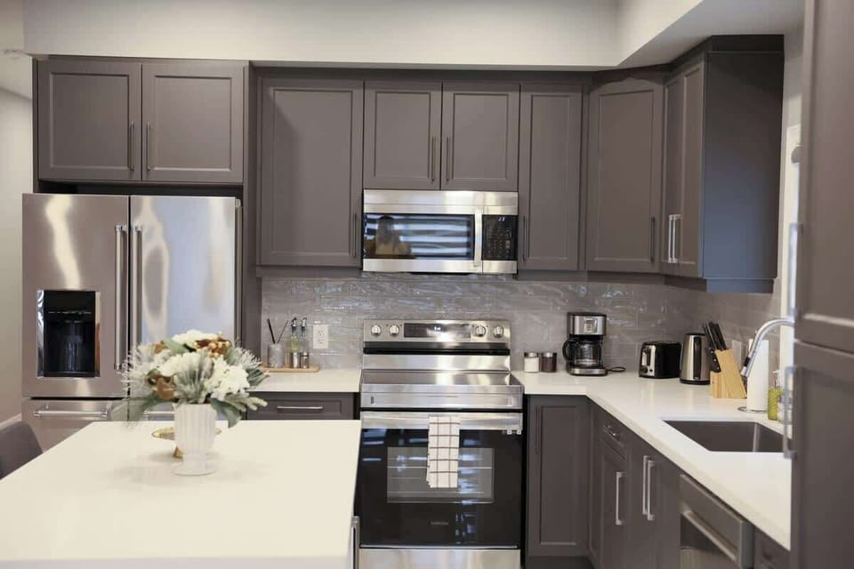 Modern kitchen featuring sleek stainless steel appliances, spacious white island, ample dark cabinetry, polished countertops, and decorative flowers.
