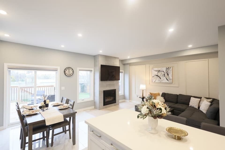 Spacious open-concept layout with elegant dining area, cozy sectional, modern fireplace, wall-mounted TV, floral centerpiece, and large sunlit windows.