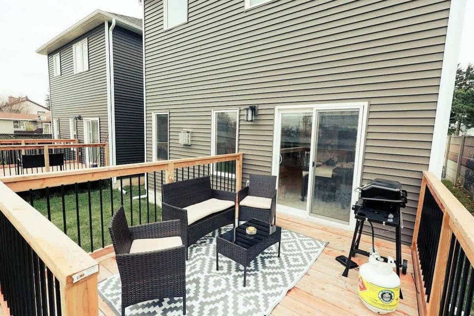 Charming private deck with cozy wicker seating, plush cushions, elegant coffee table, patterned rug, safety railing, and a sleek BBQ grill for outdoor dining.