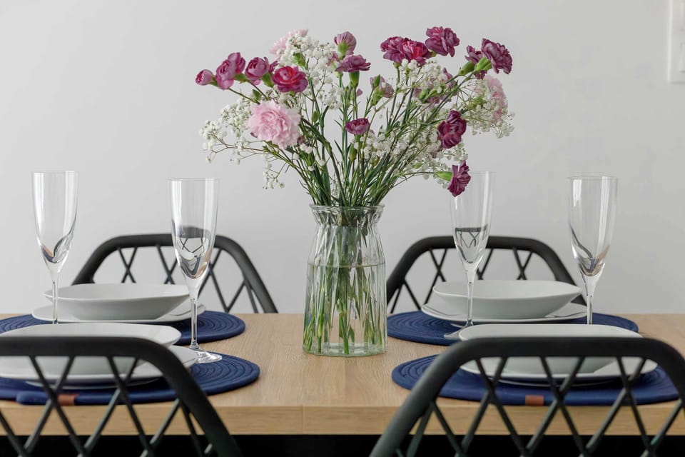 A beautifully set wooden dining table features elegant black patterned chairs, white plates, and polished silverware. A glass vase filled with fresh pink and white flowers serves as a vibrant centerpiece.
