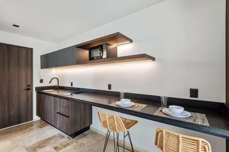 Open kitchenette with induction cooktop, microwave & matte black fixtures. Features dark wood bar & artisanal stools, perfect for preparing breakfast or enjoying morning coffee in comfort.