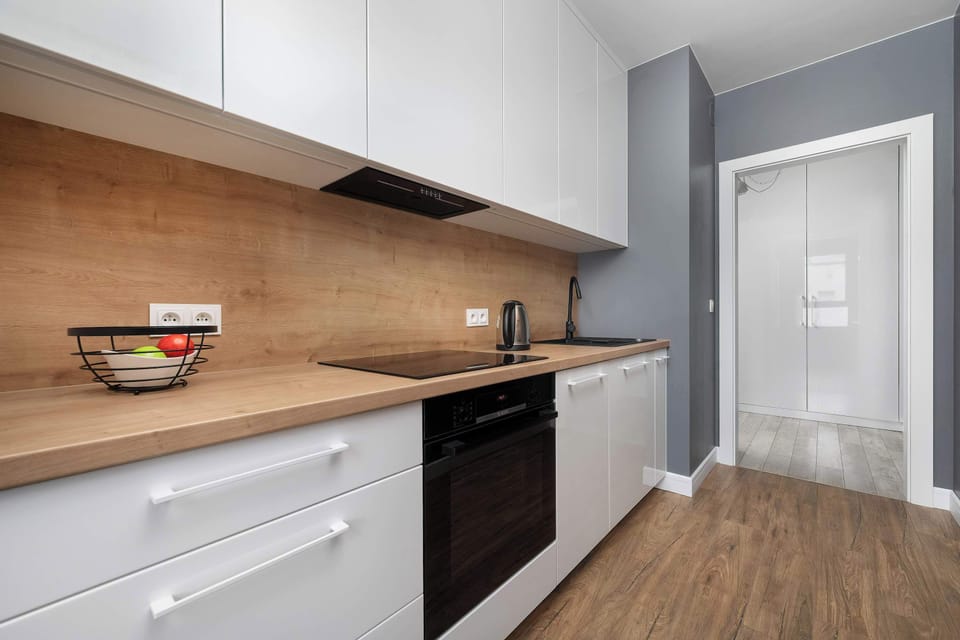 A long kitchen counter features a warm wood-toned surface contrasted with minimalist white lower cabinetry. The open layout extends toward the entryway, highlighting the apartment's smooth, open floor plan.