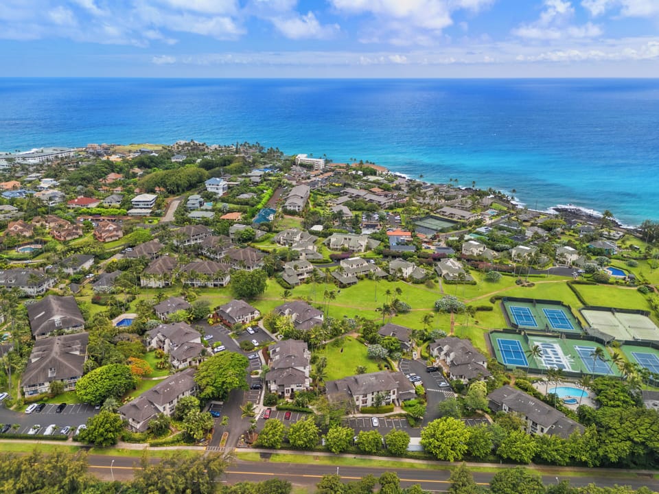 Aerial view of a stunning coastal neighborhood surrounded by turquoise ocean waters, lush greenery, and tennis courts. The property is located in a vibrant tropical community near pristine beaches.