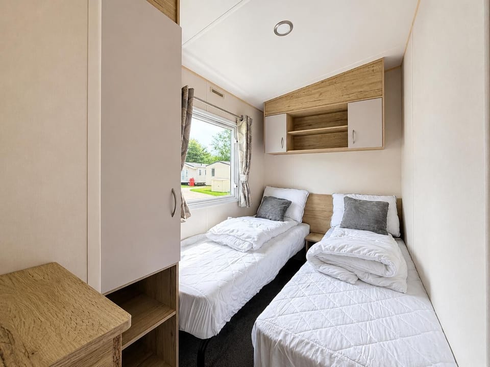 Twin bedroom with white quilted bedding, grey accent pillows, wood-paneled headboard, and overhead storage cabinets.