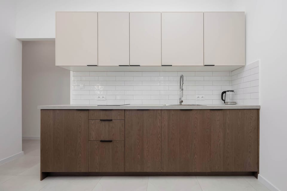 A minimalist kitchenette lines the wall, showing wood-grained lower cabinets paired with smooth, off-white upper cupboards. The setup includes a white tiled backsplash and a stainless steel sink.