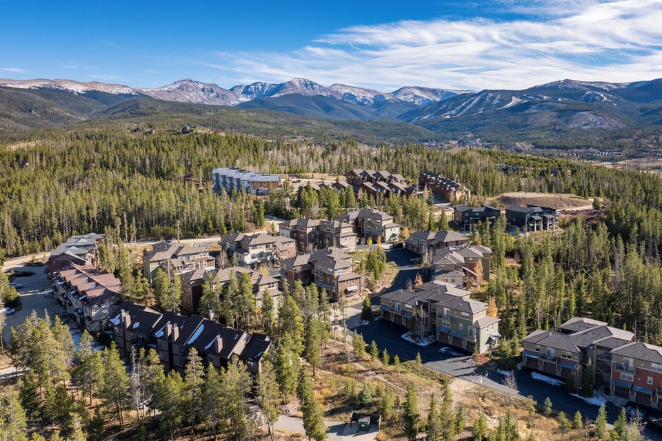 Aerial view of mountain property nestled in evergreen forest with dramatic snow-capped peaks framing the horizon and expansive wilderness beyond.