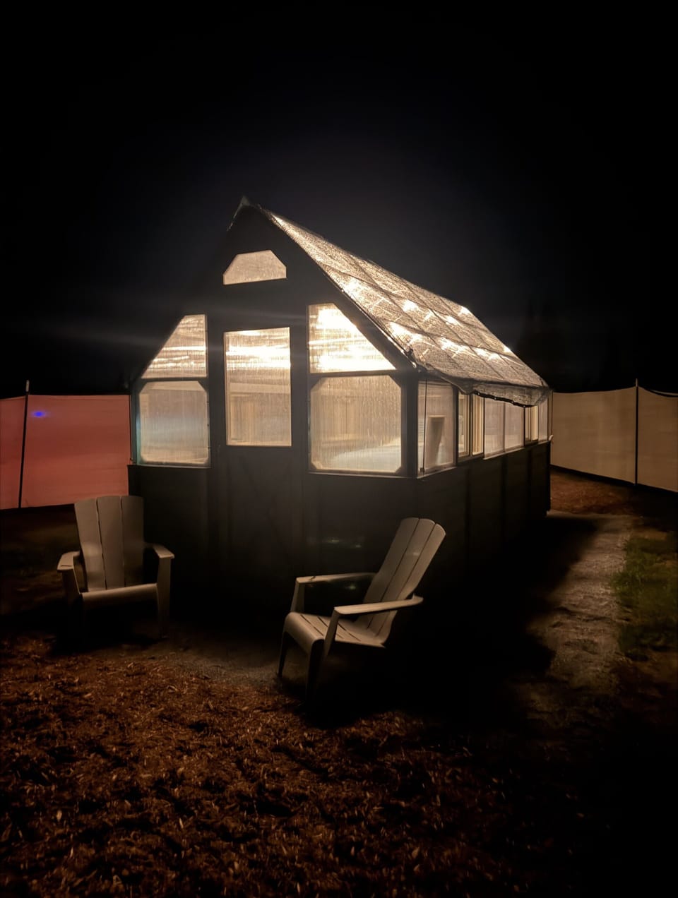 Cozy illuminated greenhouse retreat with Adirondack seating at night