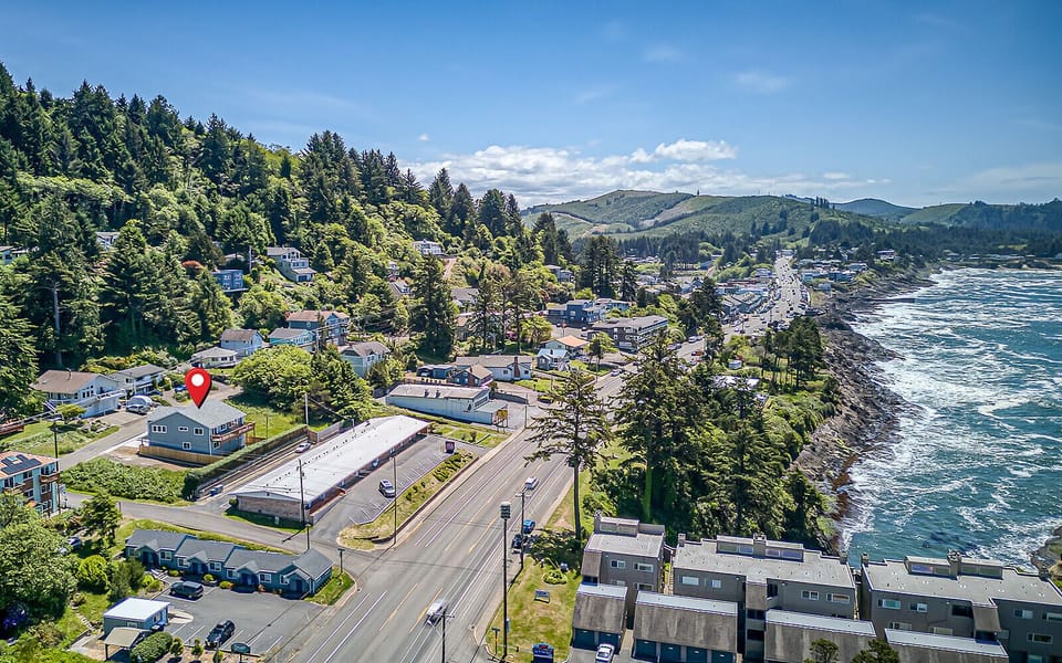 Aerial view of a coastal town with lush forested hillsides, ocean waves, and winding streets—a tranquil setting close to nature's beauty.