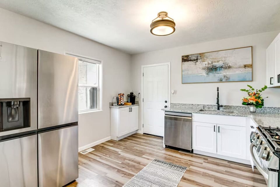 The full kitchen layout — stainless steel fridge, dishwasher, gas range, white shaker cabinets, and granite counters, all on warm hardwood floors. Fully stocked with cookware, dishware, and everything you need. No grocery trip anxiety here.