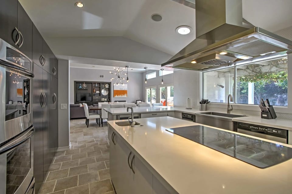 Modern chef's kitchen with a large waterfall-edge island, induction cooktop, stainless hood, double wall ovens, and a window over the sink.