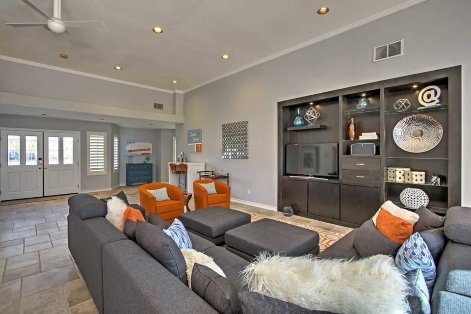 Comfortable living room with a large gray sectional, ottomans, orange accent chairs, built-in entertainment center, and a bright entryway beyond.