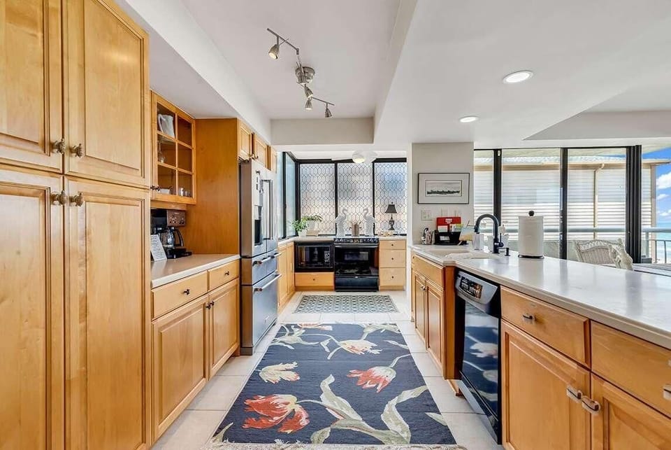Maple cabinetry lines the galley kitchen, with black appliances and a floral runner on the tiled floor.