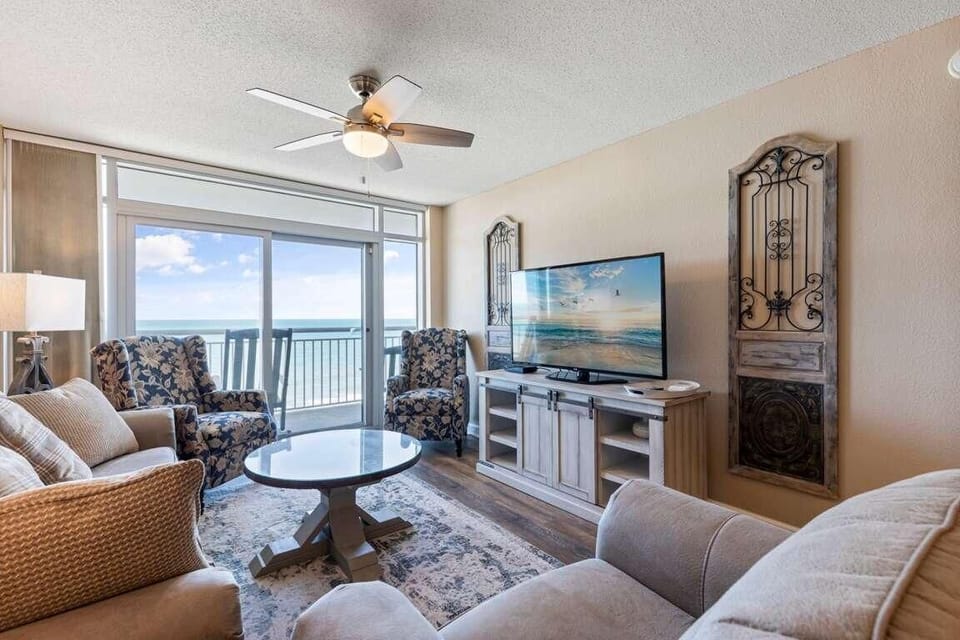 Living room with floral armchairs, wicker sofa, a round wooden coffee table, and ocean views through glass doors.