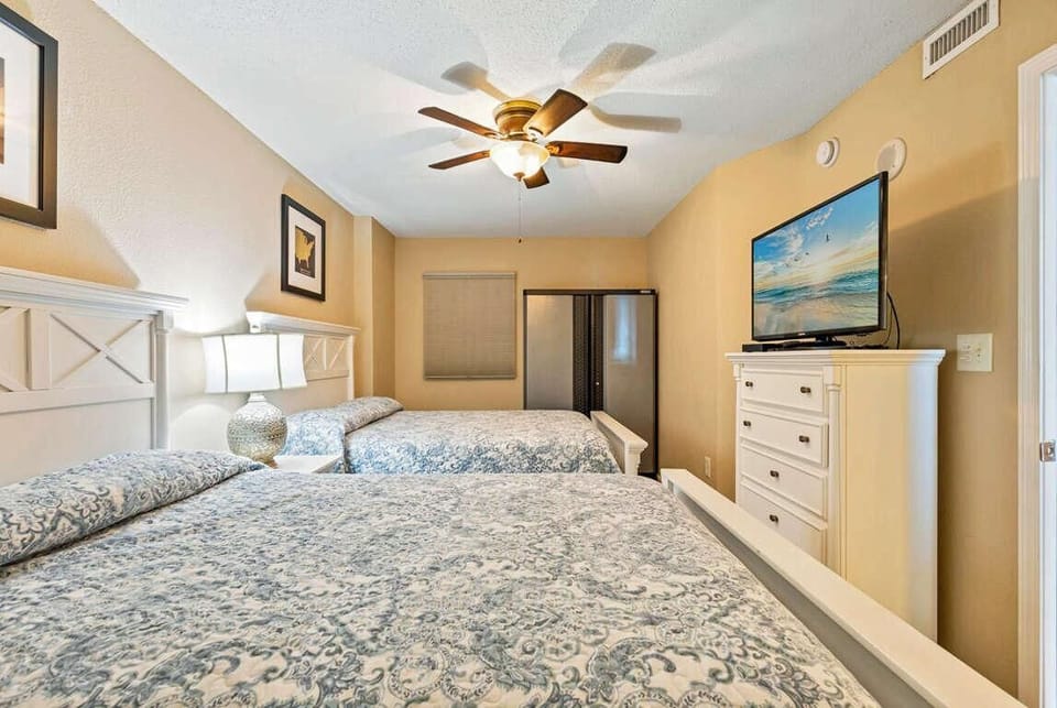 Two white wooden beds with patterned blue bedding face a dresser topped with a flat-screen TV.