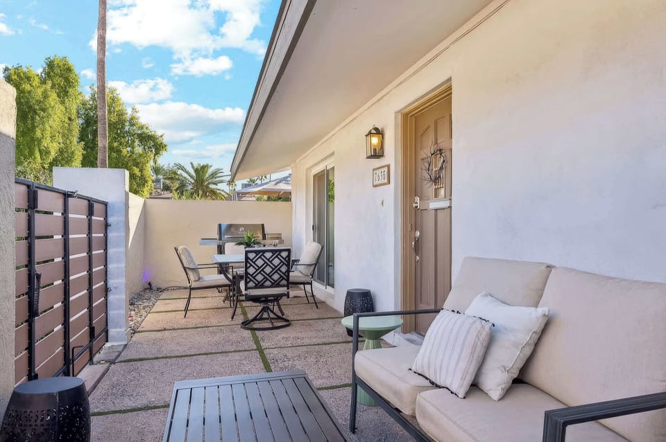 Private patio with lounge seating, a four-seat dining set, gas grill, retractable umbrella, and gated entry framed by swaying palms and Arizona skies.