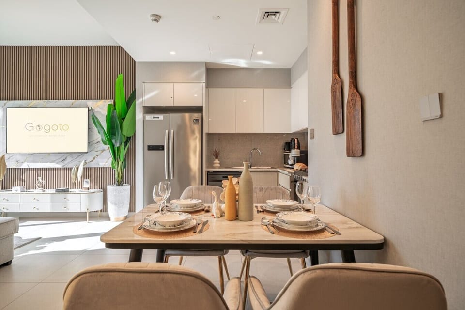 Bright and stylish dining area featuring comfortable seating for four, a large Smart TV, decorative plants, and a fully equipped kitchen with a spacious double-door refrigerator—perfect for enjoying meals and spending time together.
