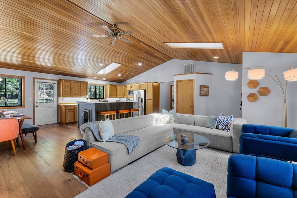 Unwind in your open-concept living space featuring vaulted wood ceilings, modern furnishings in calming blues and grays, and a fully-equipped kitchen. The warm wood tones and natural light create a welcoming retreat for your stay.