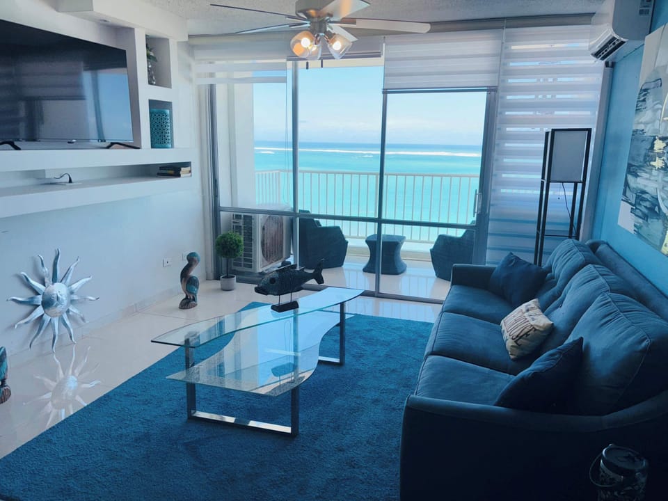 Bright coastal living room with floor-to-ceiling ocean views, modern blue accents, and a cozy sofa. Perfect for relaxing while soaking in the beauty of the sea.