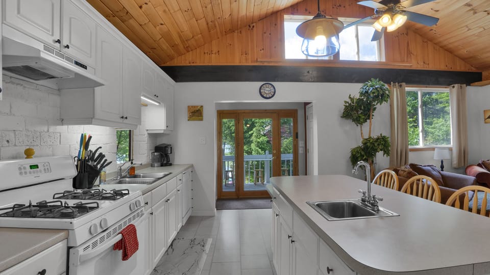 Bright, modern kitchen with white cabinetry, wood ceiling accents, and a view of the outdoor deck and forest.