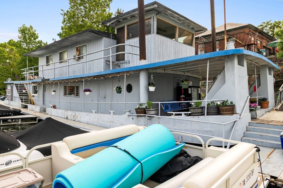 Houseboat front at the marina with hanging planters, porthole window, and pontoon boats in the foreground.