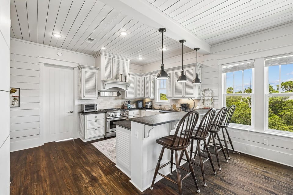 Your bright coastal kitchen features modern appliances and a generous island for gathering, framed by windows overlooking natural greenery.