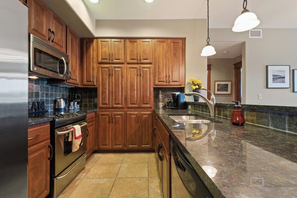 A well-appointed galley kitchen featuring extensive wooden cabinetry, polished dark granite countertops with a breakfast bar, stainless steel appliances, and a warm, neutral tile floor.