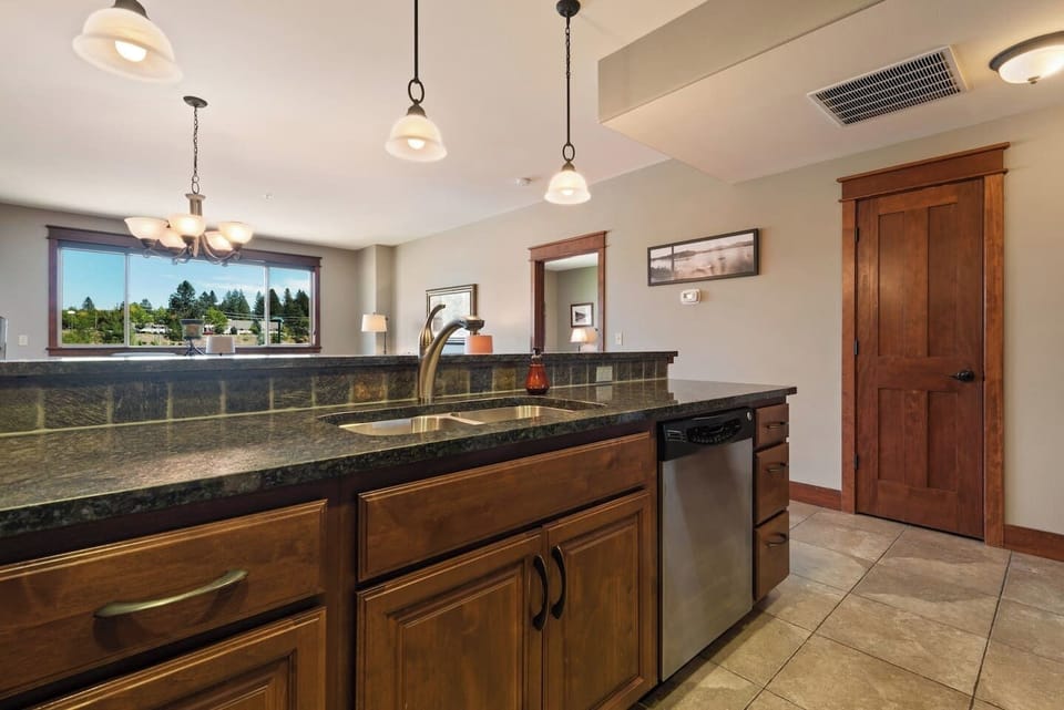 A perspective from behind the kitchen island counter showcasing a double-basin stainless steel sink and a built-in dishwasher, with neutral tile flooring below and wood-paneled doors nearby.