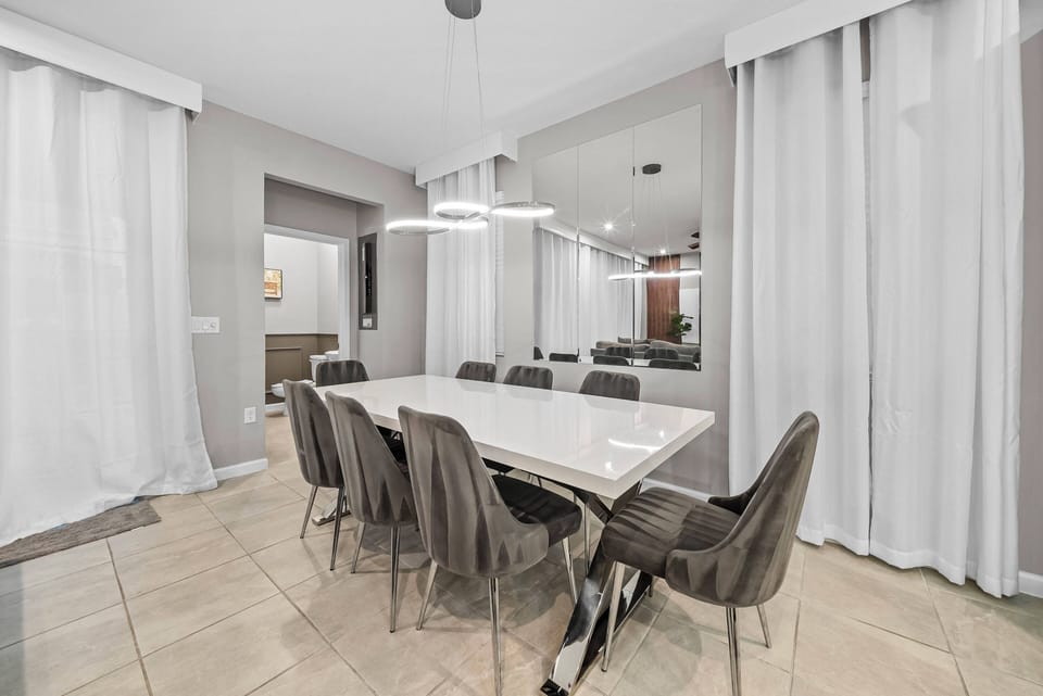 Sleek and contemporary, this dining area features a spacious white table surrounded by stylish gray chairs, perfect for gatherings. The soft natural light filters through elegant curtains, enhancing the inviting atmosphere.