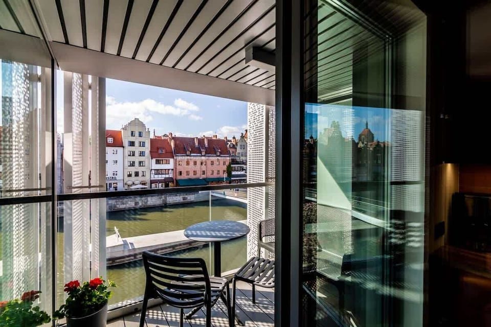 Balkon zachwyca widokiem na rzekę i okoliczną zabudowę, a komplet mebli pozwala wygodnie delektować się poranną kawą lub wieczornym odpoczynkiem na świeżym powietrzu.