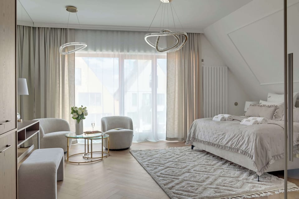 Przestronna sypialnia premium urządzona w jasnych, eleganckich barwach. Komfortowe łóżko, strefa relaksu z fotelami, duże przeszklenia oraz stylowe oświetlenie tworzą wyjątkową atmosferę sprzyjającą wypoczynkowi i regeneracji.