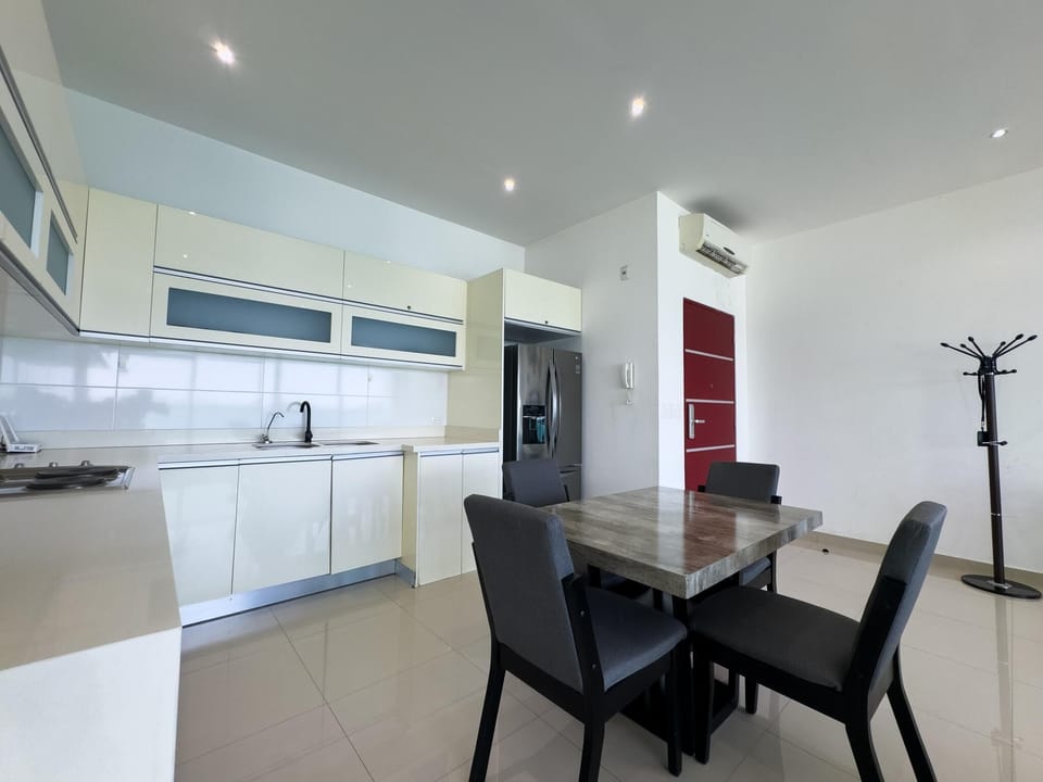 Dining table with gray upholstered chairs sits near white cabinetry and stainless steel refrigerator in kitchen area.