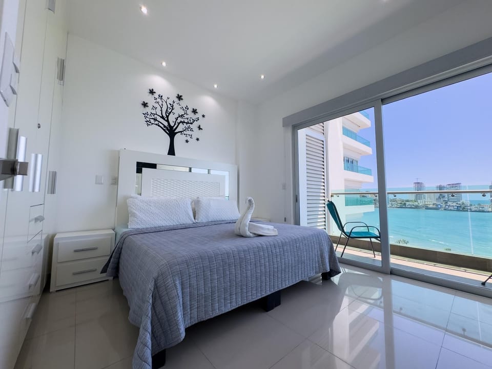 Master bedroom with glossy white tile floor, gray quilted bedspread, black tree wall art, and ocean-view balcony.