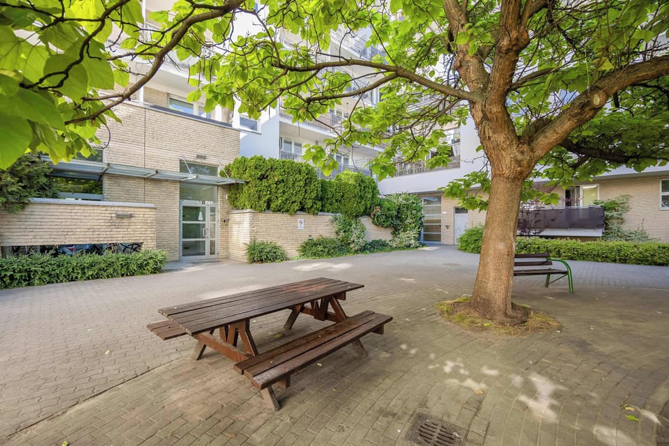 A picnic table beneath leafy trees creates a peaceful communal seating area surrounded by greenery. The courtyard offers a pleasant place to relax outdoors.
