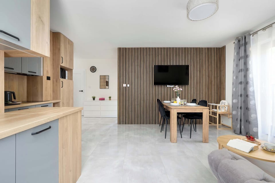 A wide view displays the living room with a slatted wood accent wall and a wall-mounted TV. The dining table and kitchen setup complete the space.
