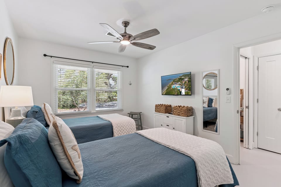 Unwind in this bright, coastal-inspired guest room with two plush beds, a ceiling fan for comfort, and charming touches like woven baskets and natural light from the windows.
