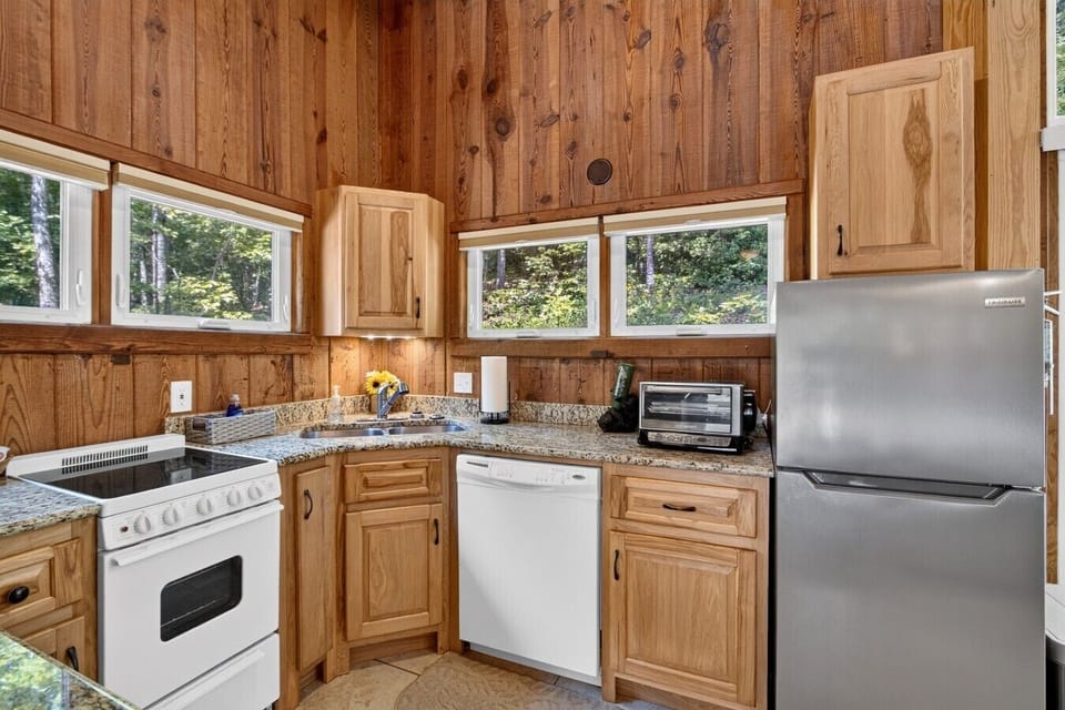 Full kitchen with granite counters, dishwasher, stove, fridge, and toaster oven — windows on two sides look into the trees.