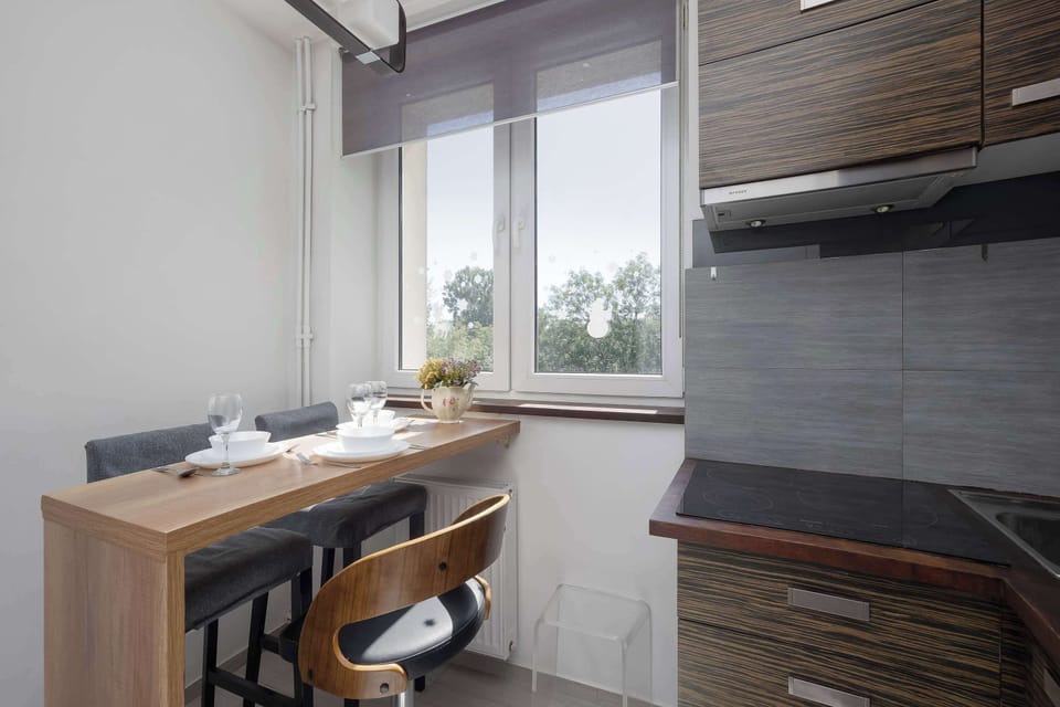A modern kitchen with wooden cabinets, black countertops, and a dining table by the window. Large windows make the space bright and inviting.