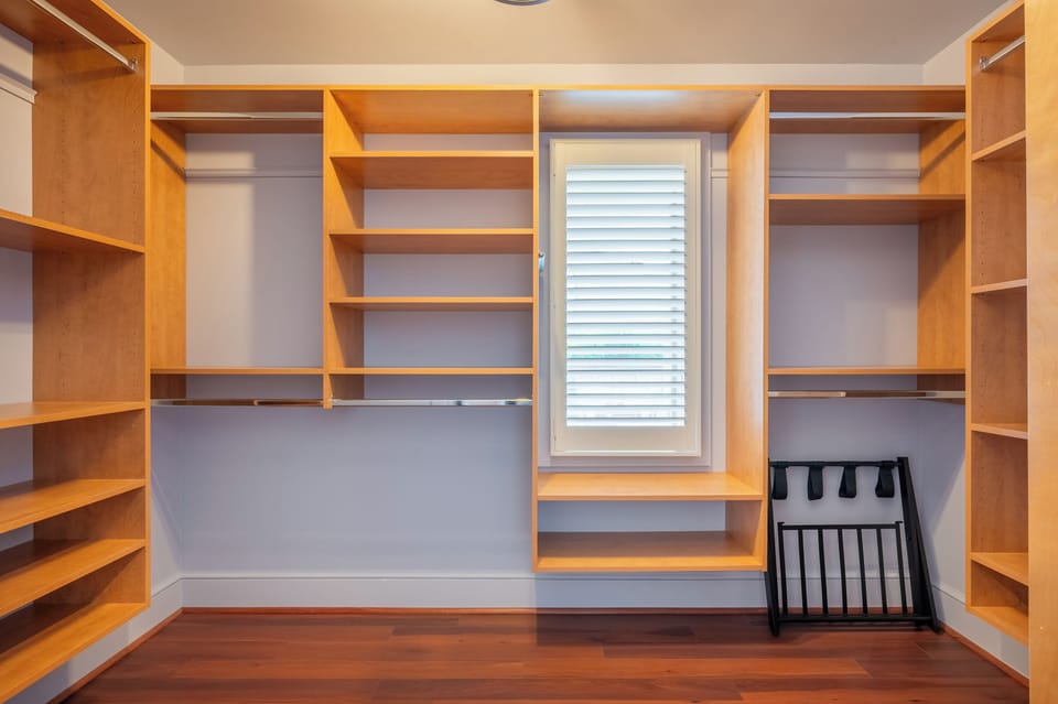 An empty walk-in closet with custom wood shelving, clothing rods, and a small shuttered window.