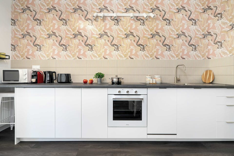 A sleek, contemporary kitchenette showcases white handleless cabinetry paired with a dark countertop and a decorative patterned backsplash. Built-in appliances like an oven and stove are neatly integrated into the workspace.